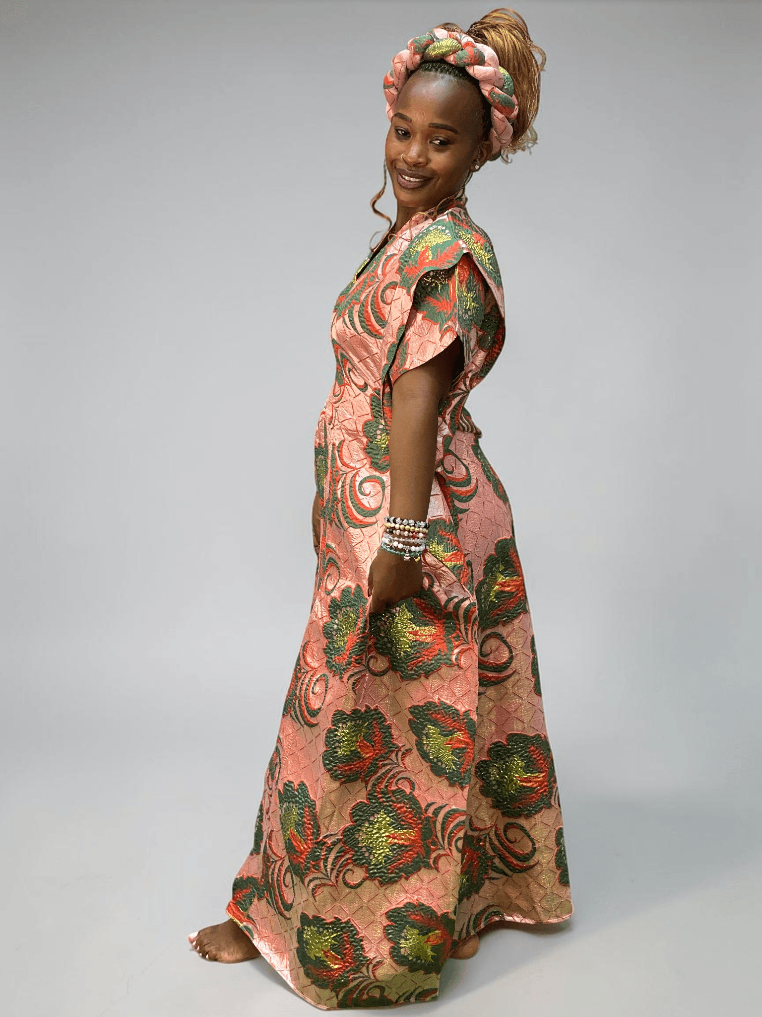 Coral pink and green floral brocade gown with woven textured pattern and adjustable waist ties on grey studio background