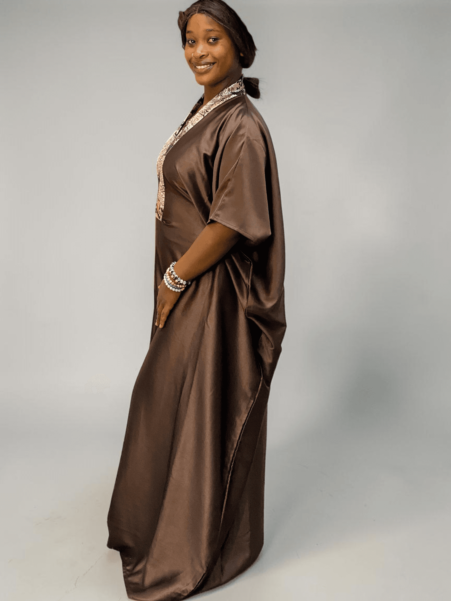Chocolate brown satin boubou gown with brocade neckline trim and adjustable waist ties on grey studio background