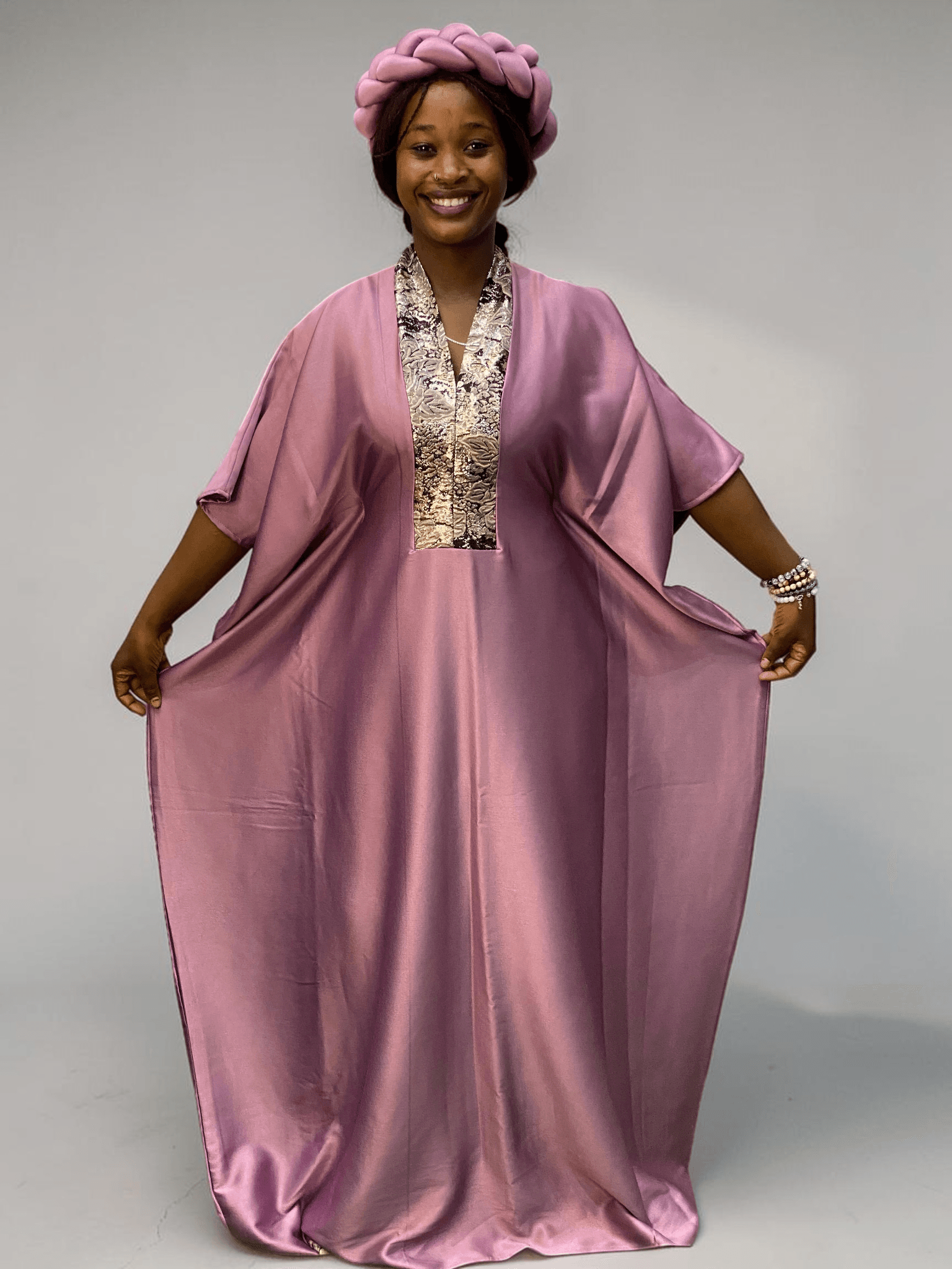 Blush pink satin boubou gown with brocade neckline trim and adjustable waist ties on grey studio background