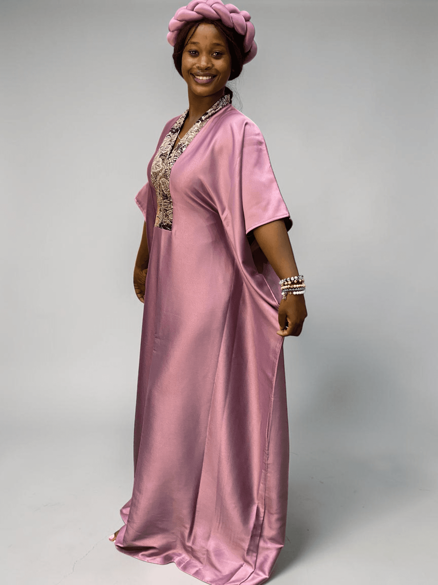 Blush pink satin boubou gown with brocade neckline trim and adjustable waist ties on grey studio background